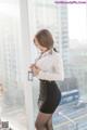 A woman in a white shirt and black skirt standing by a window.