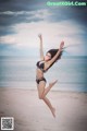 A woman in a black bikini jumping in the air on a beach.